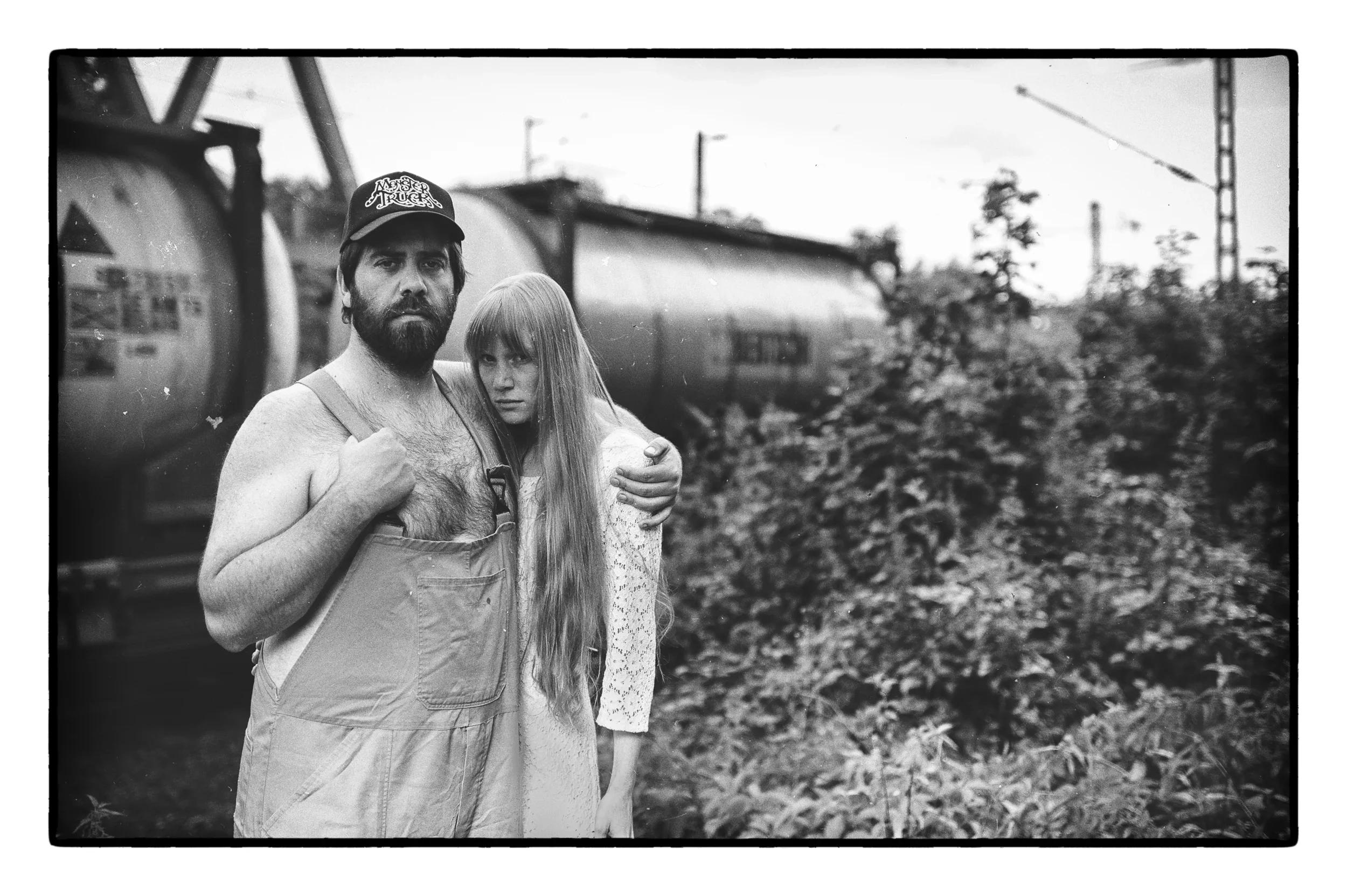 Wide black and white composition of a couple standing in front of industrial tanks in the woods.